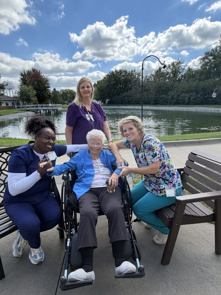 elderly woman in wheelchair smiling outside with three female nurses surrounding her.